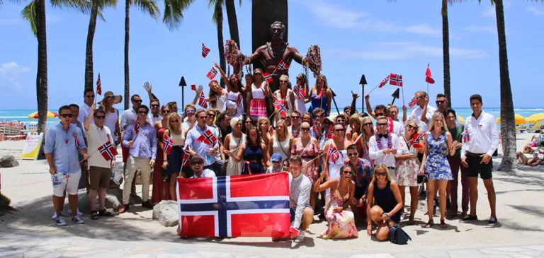 Stor gruppe med blide mennesker vifter med norske flagg på en strand