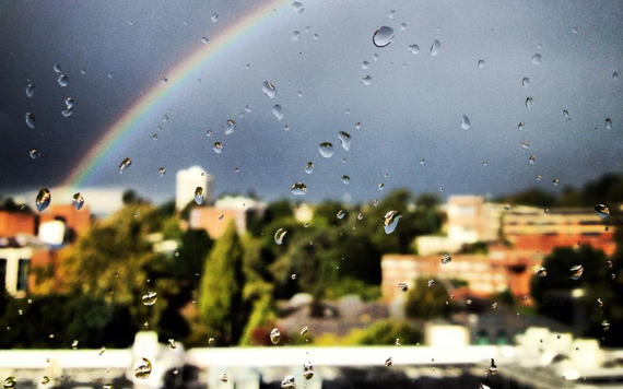Regnbue bak en by sett gjennom vindu med regn på
