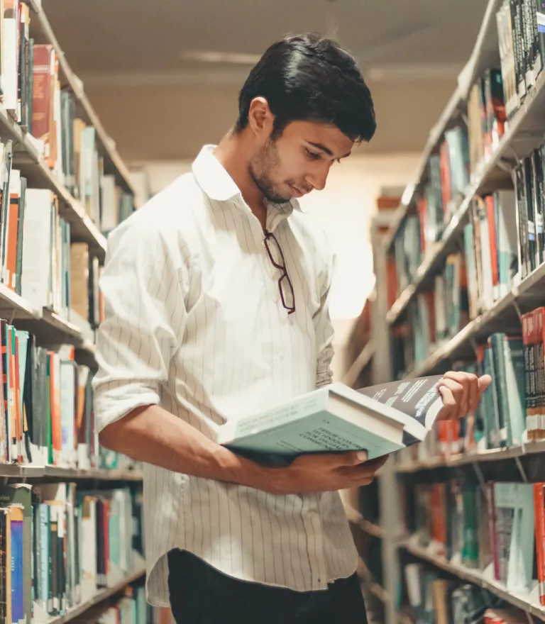 Ung mann i hvit skjorte står mellom bokhyller og leser en bok