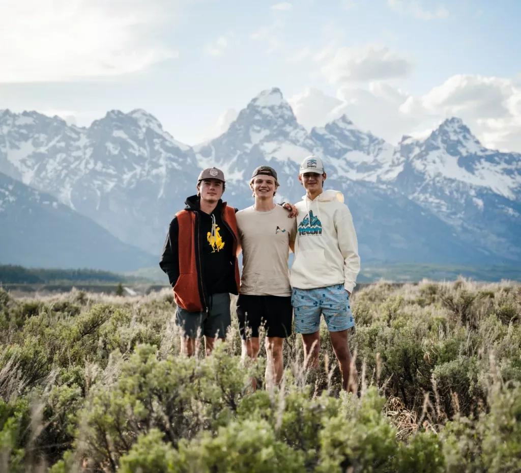 Tre smilende, unge menn står i gress foran en fjellkjede
