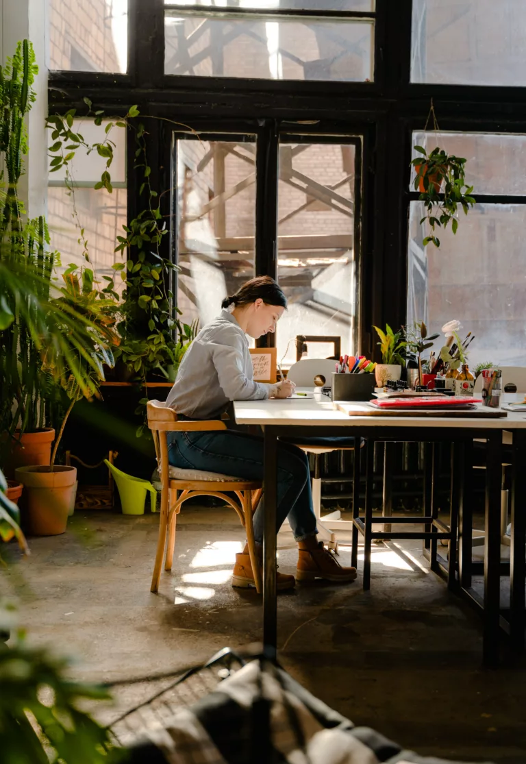 Kvinne sitter i stol foran et bord og jobber, med planter rundt seg