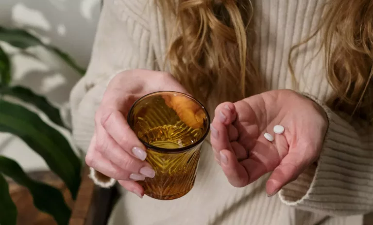 Kvinne med langt blondt hår har et glass vann i en hånd, og to piller i den andre hånden