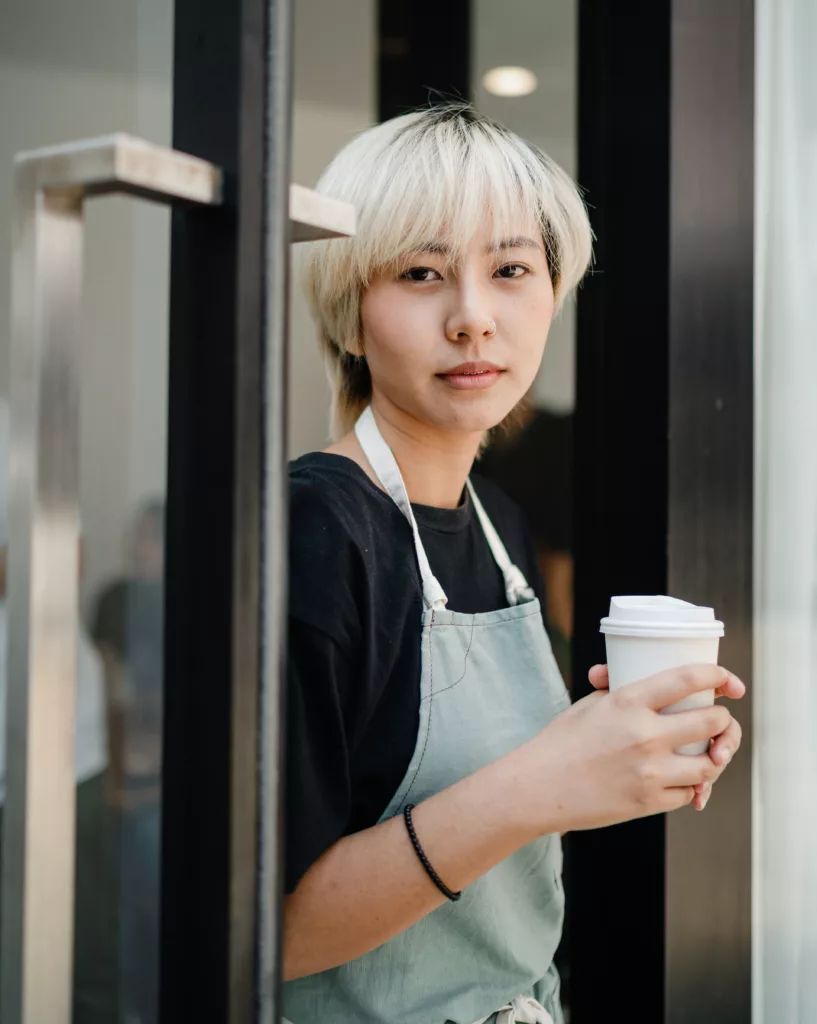 Kvinne med lyst, kort hår og servitør-uniform holder en kaffe