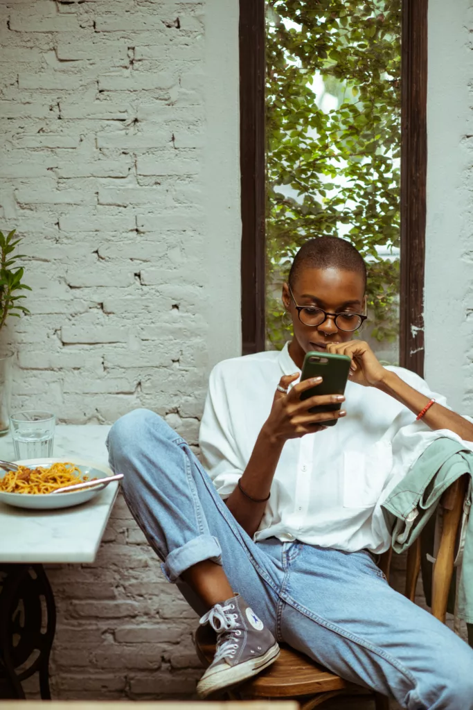 Ung kvinne med kort hår og jeans sitter på en stol og leser noe på telefonen sin