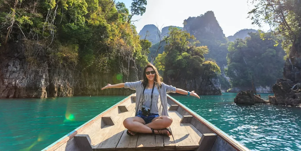 Ung kvinne med solbriller sitter med utstrakte armer i en trebåt