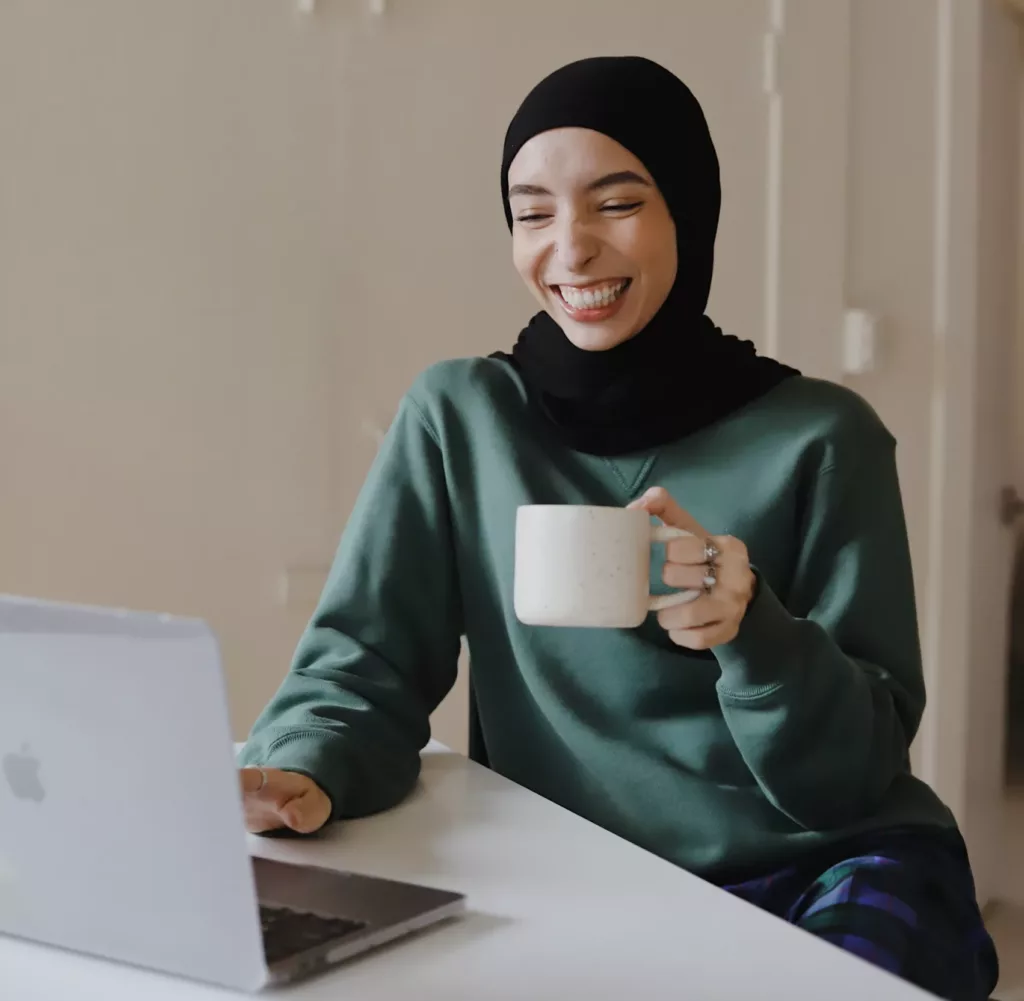 Ung, smilende kvinne med hijab sitter ved en PC mens hun holder en kopp