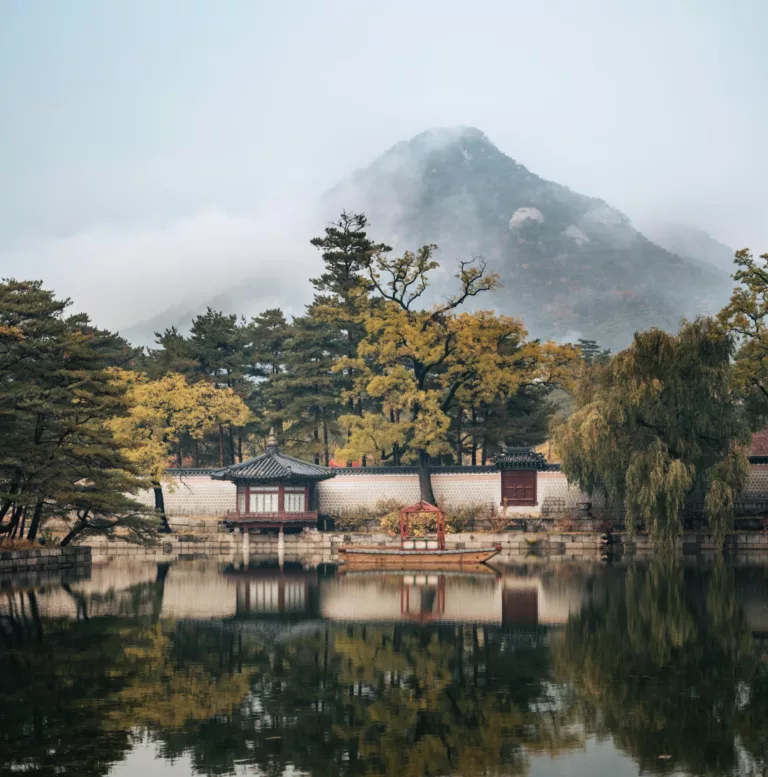 Koreansk hus ved vannet. Et fjell stikker opp bak trærne i bakgrunnen.