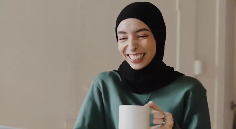 Ung, smilende kvinne med hijab sitter ved en PC mens hun holder en kopp