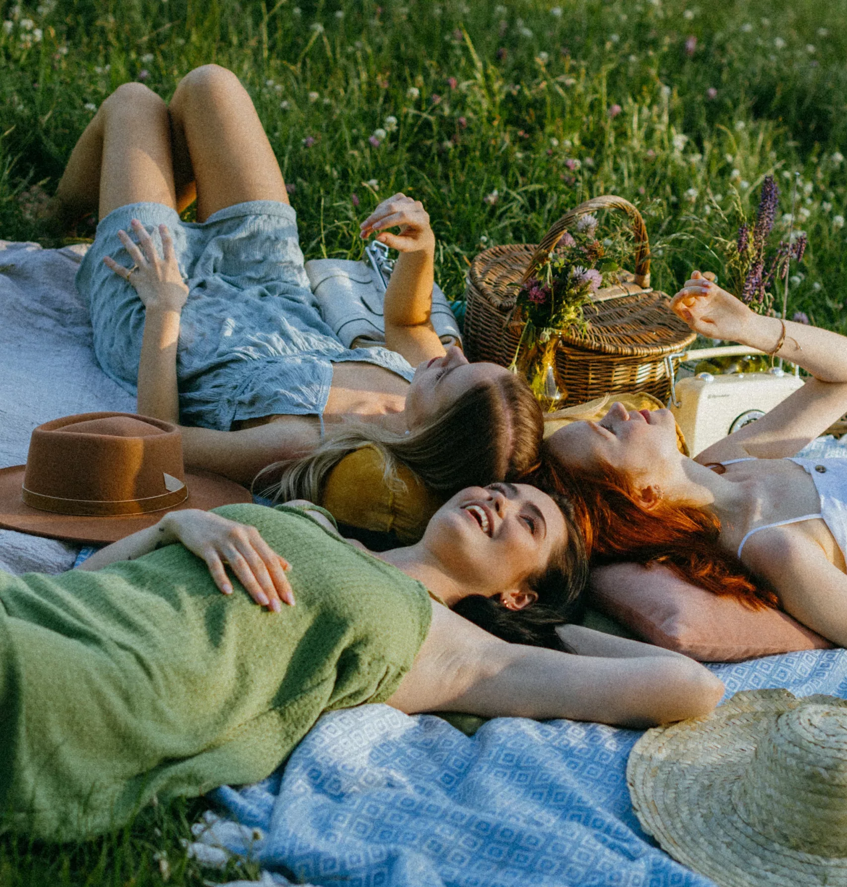 Tre unge kvinner ligger på ryggen på et piknikteppe i gresset