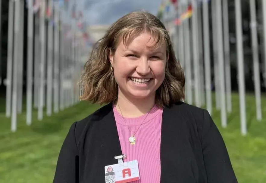 Ung kvinne med svart jakke og rosa topp smiler foran to rekker med flagg