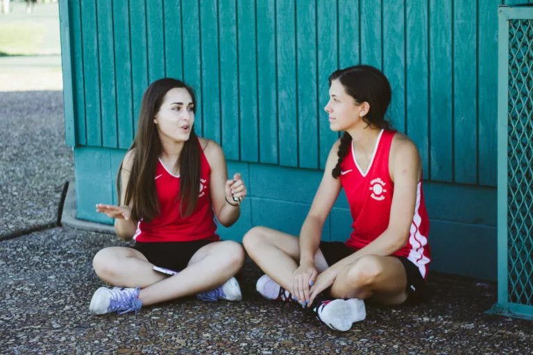 To jenter i rødt sportstøy sitter på bakken ved en grønn vegg og snakker