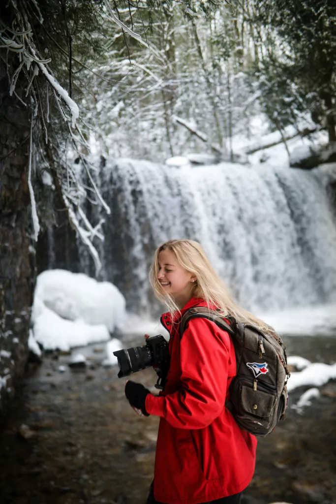 En ung, blond kvinne med rød anorakk og grønn ryggsekk står foran en foss i Ontario, Canada