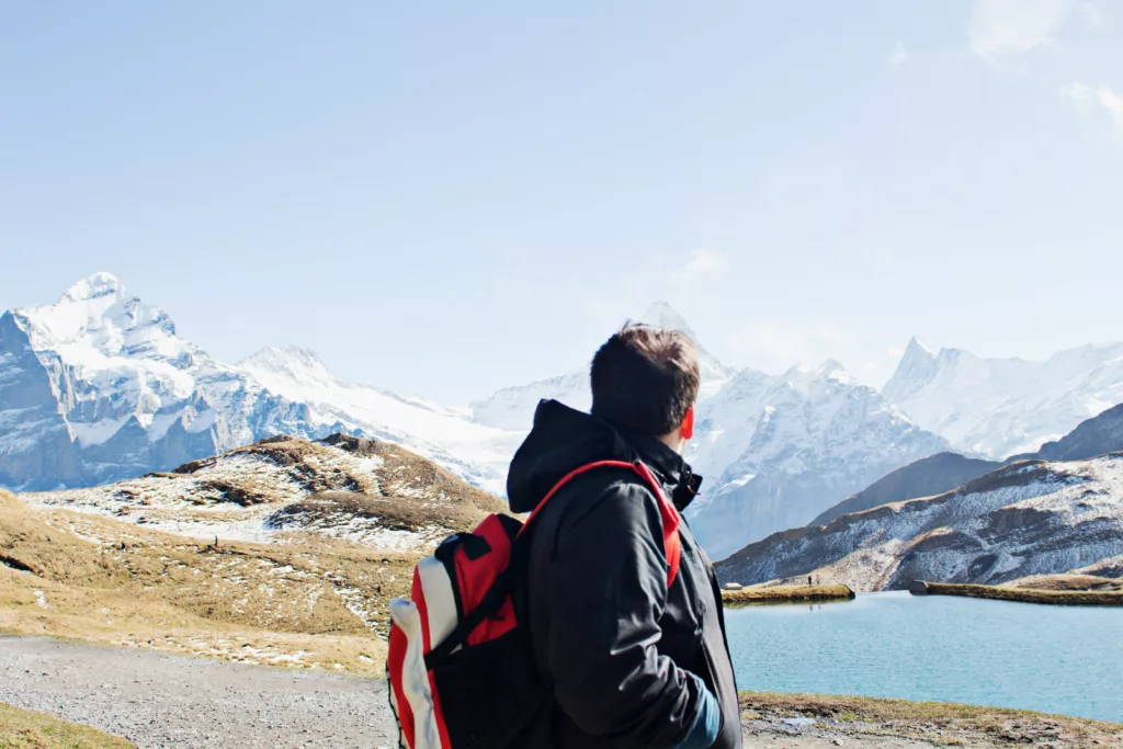 En ung mann med svart jakke og rød ryggsekk ser utover alpene i Sveits