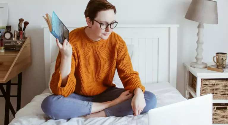 Kvinne i oransje genser sitter på en seng og jobber med PC