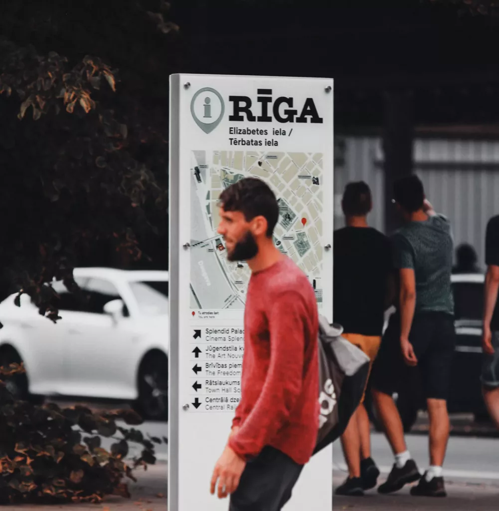 En mann med rød genser, svart bukse og røde sko går foran et informasjonsskilt med "Riga" i store bokstaver.