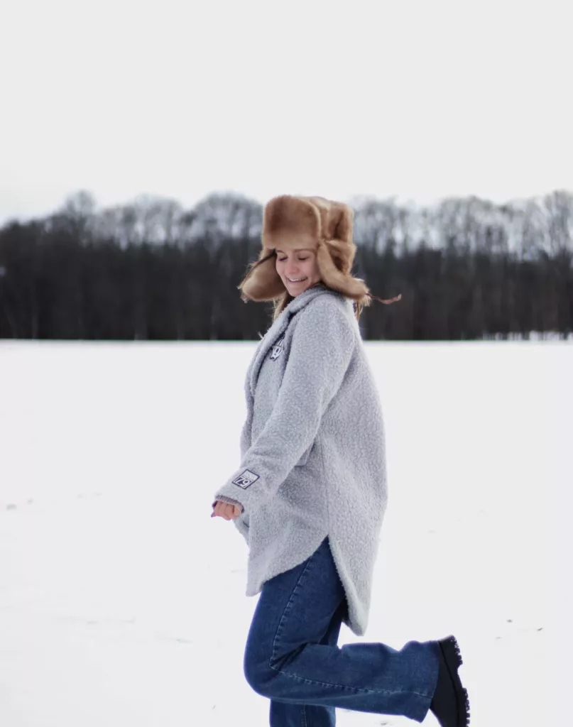 Ung kvinne med stor lue, grå jakke og jeans står i et landskap med snø