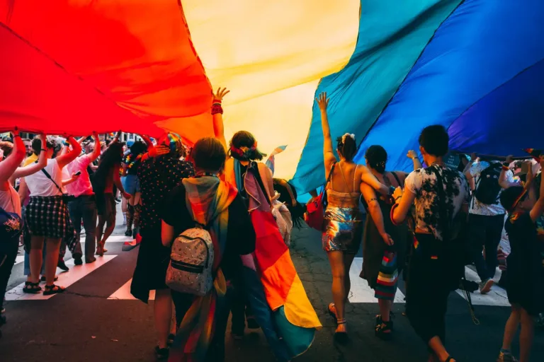 Folk går under et stort Pride-flagg i en parade